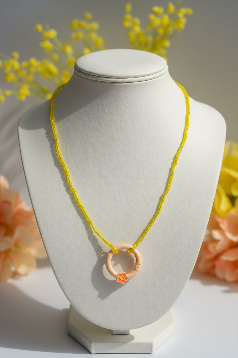 A yellow beaded necklace with a circular peach pendant featuring a small orange flower accent, displayed on a neutral gray background.