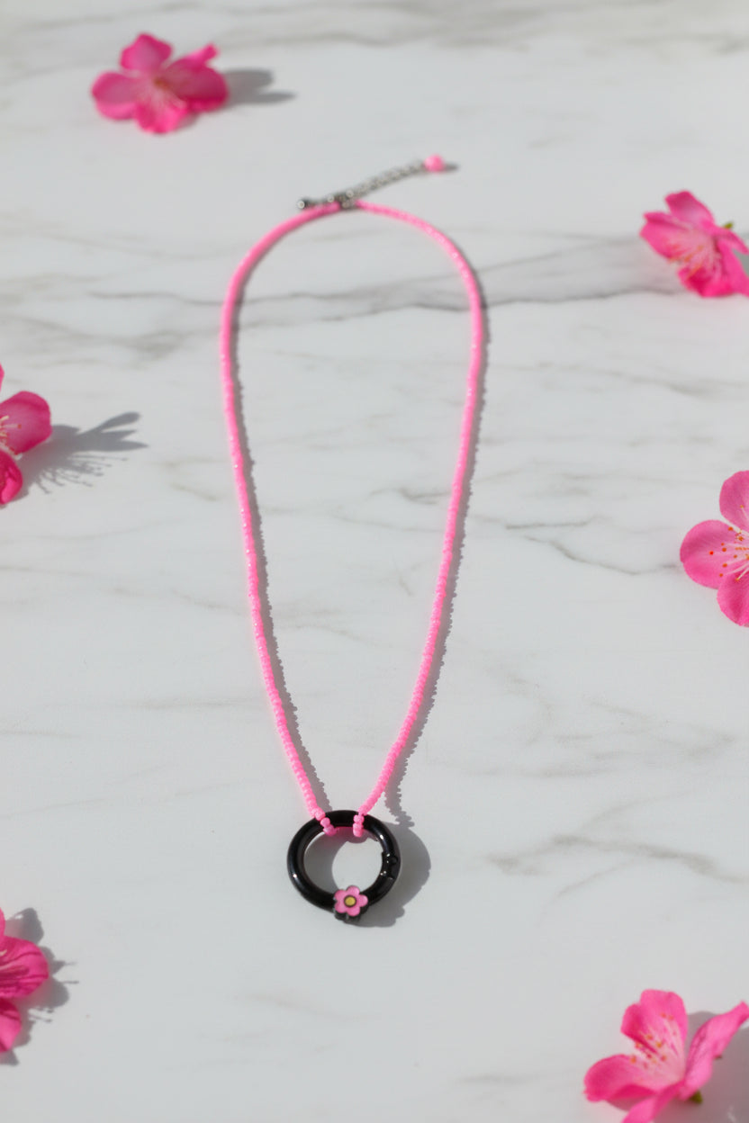 A pink beaded necklace with a black circular ring pendant featuring a small pink flower detail, displayed against a neutral gray background.