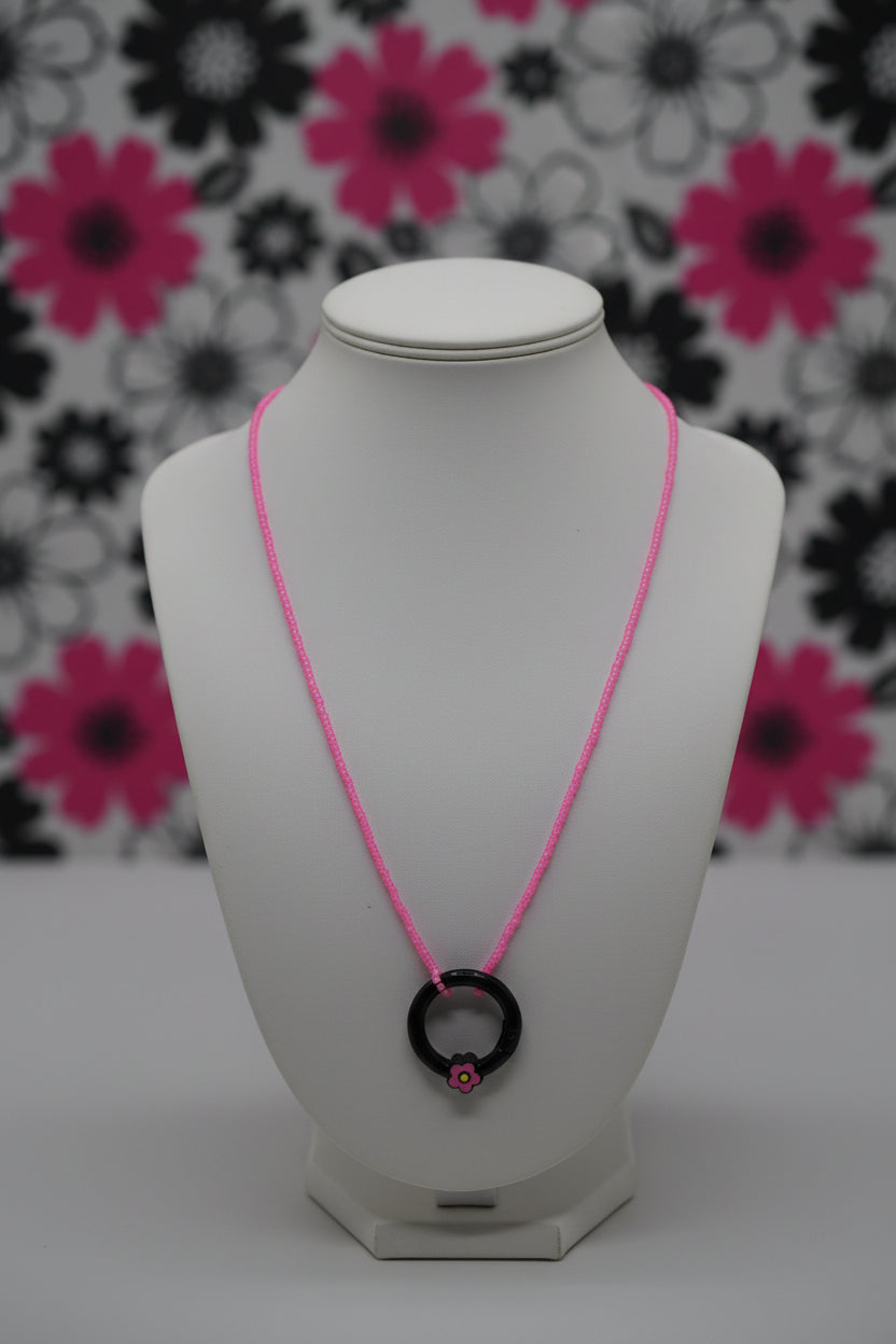 A pink beaded necklace with a black ring pendant featuring a small pink flower accent, displayed against a neutral gray background.