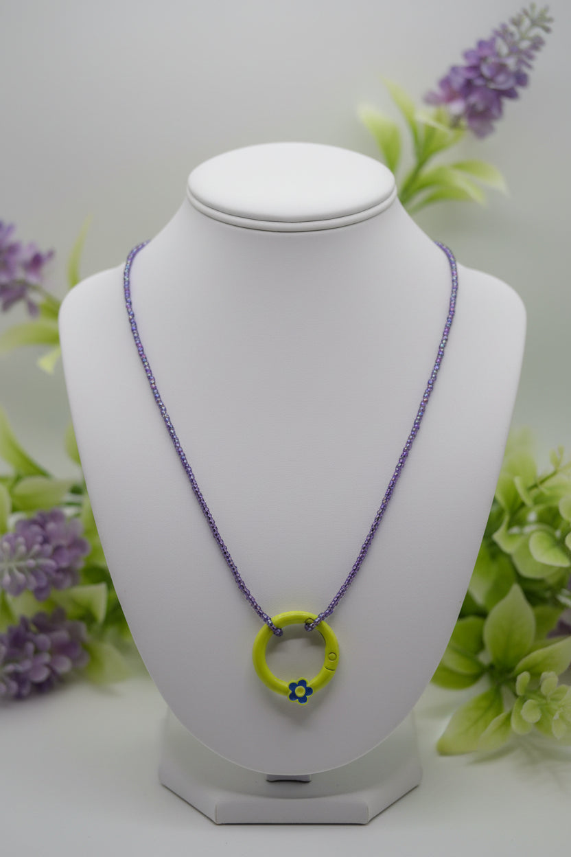 A handmade beaded necklace with a lime green circular pendant featuring a small blue and yellow flower charm, on a light gray background.