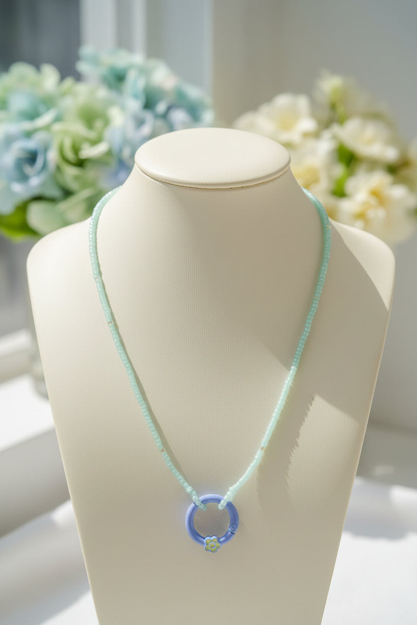 A delicate necklace with a light mint green beaded chain and a light blue circular pendant, styled on a textured white fabric with small purple flowers.
