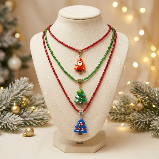 Three beaded Christmas tree necklaces displayed on a black velvet jewelry bust. The necklaces feature red, green, and red-orange beaded strands with tree pendants in orange, green, and blue, accented with small bells.