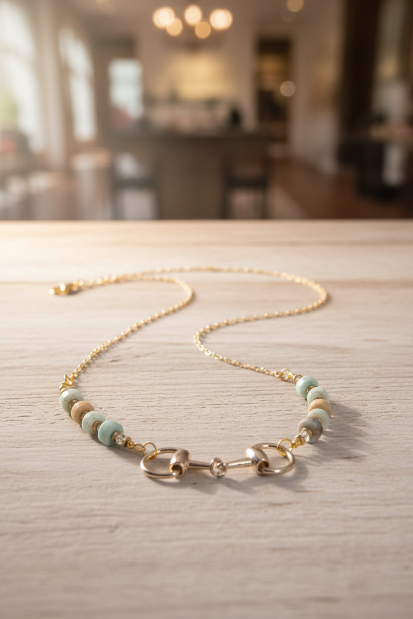 A gold-tone chain necklace displayed on a black velvet jewelry bust. The necklace features a central horse bit clasp flanked by alternating white and pale blue beads with small gold spacer details.