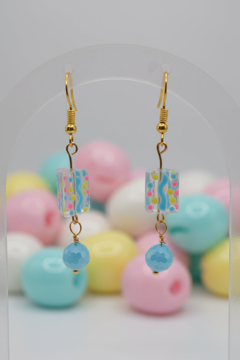 A pair of handmade dangle earrings with gold-tone fishhook wires, patterned rectangular beads, and light blue round beads, displayed on a clear acrylic stand against a neutral gray background.