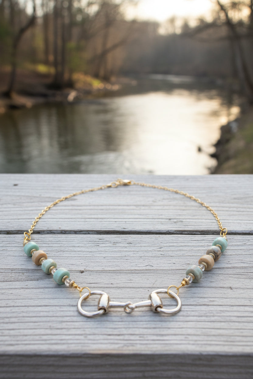 A gold-tone chain necklace displayed on a black velvet jewelry bust. The necklace features a central horse bit clasp flanked by alternating translucent white beads and small iridescent blue-green beads.