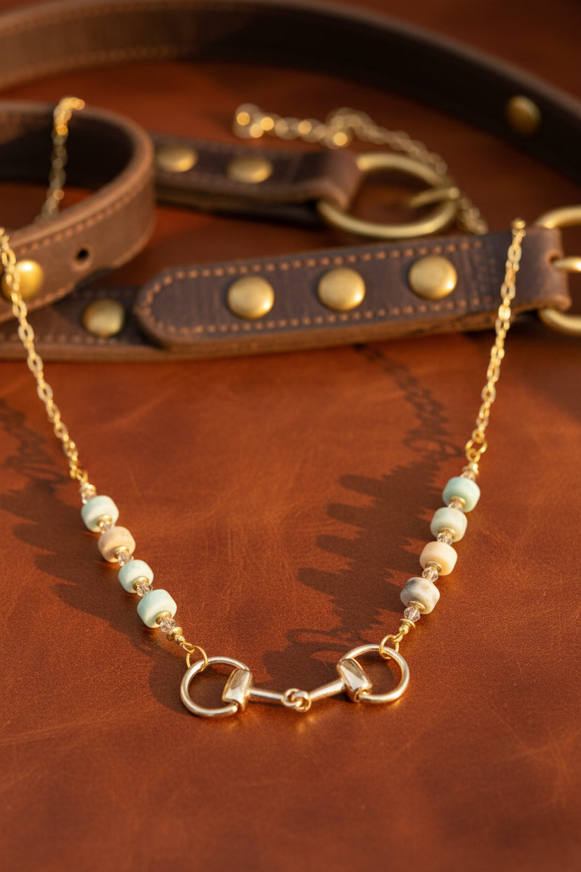 A gold-tone necklace displayed on a black velvet jewelry bust. The necklace features a horsebit clasp at the center, flanked by alternating white and pale blue beads with small gold spacer details, set against a plain white background.