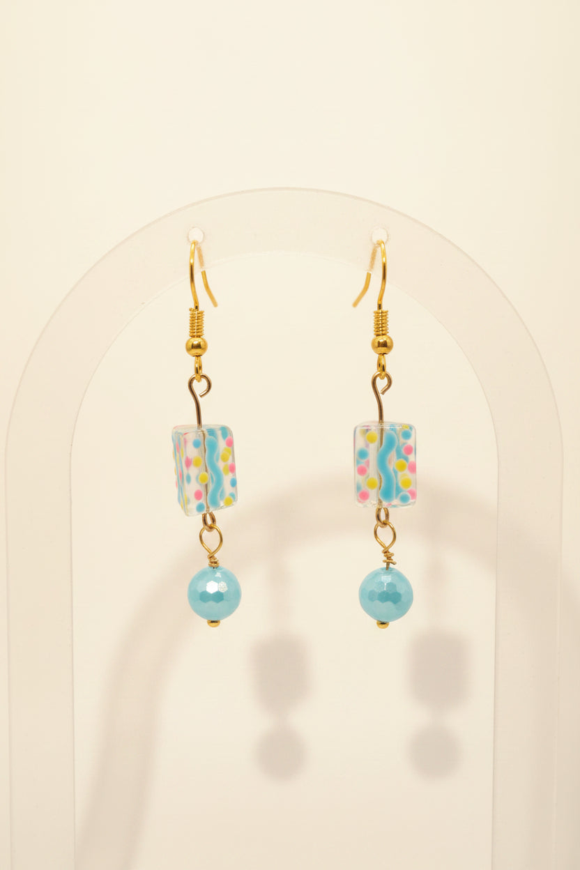 A pair of handmade dangle earrings with gold fishhook wires, patterned rectangular glass beads, and light blue glass beads, displayed on a clear acrylic stand against a neutral gray background.