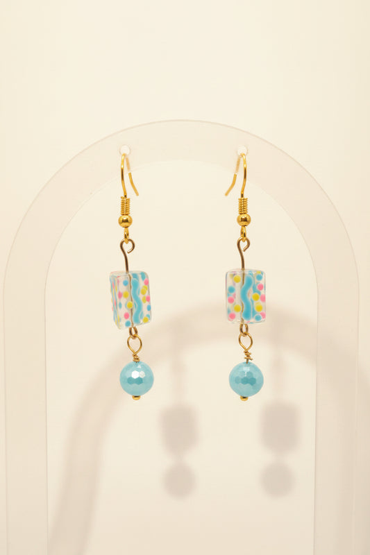 A pair of handmade dangle earrings with gold fishhook wires, patterned rectangular glass beads, and light blue glass beads, displayed on a clear acrylic stand against a neutral gray background.