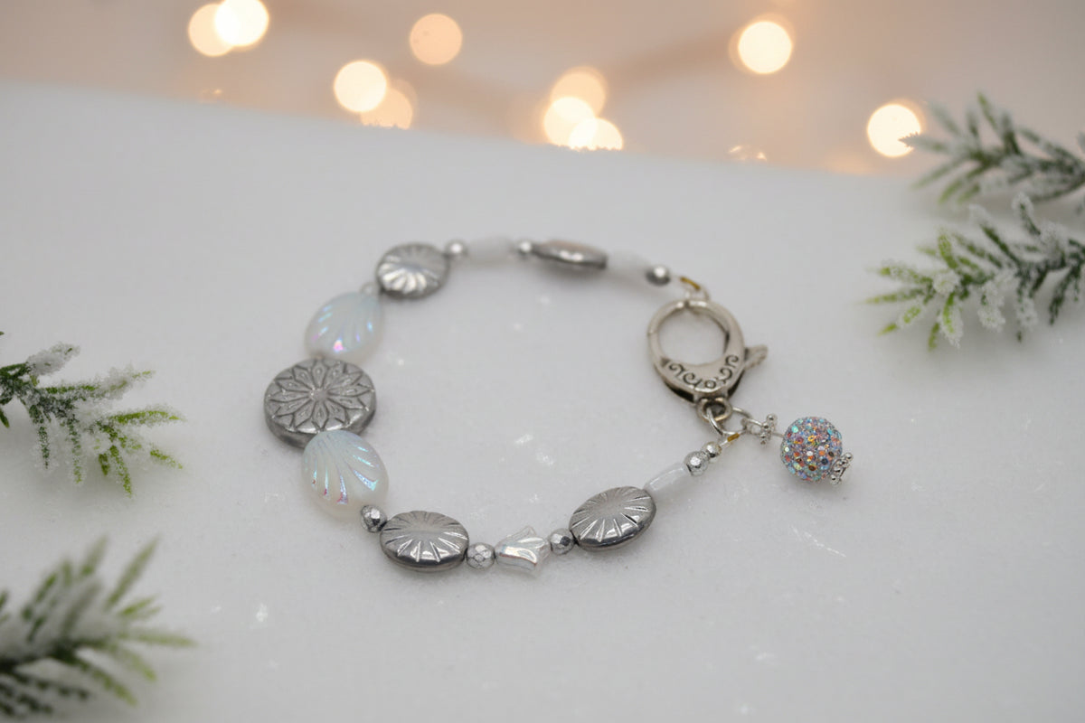 A beaded bracelet featuring silver-toned metal beads, white translucent beads, iridescent oval beads, and a small rhinestone-embellished ball charm, displayed on a neutral gray background.