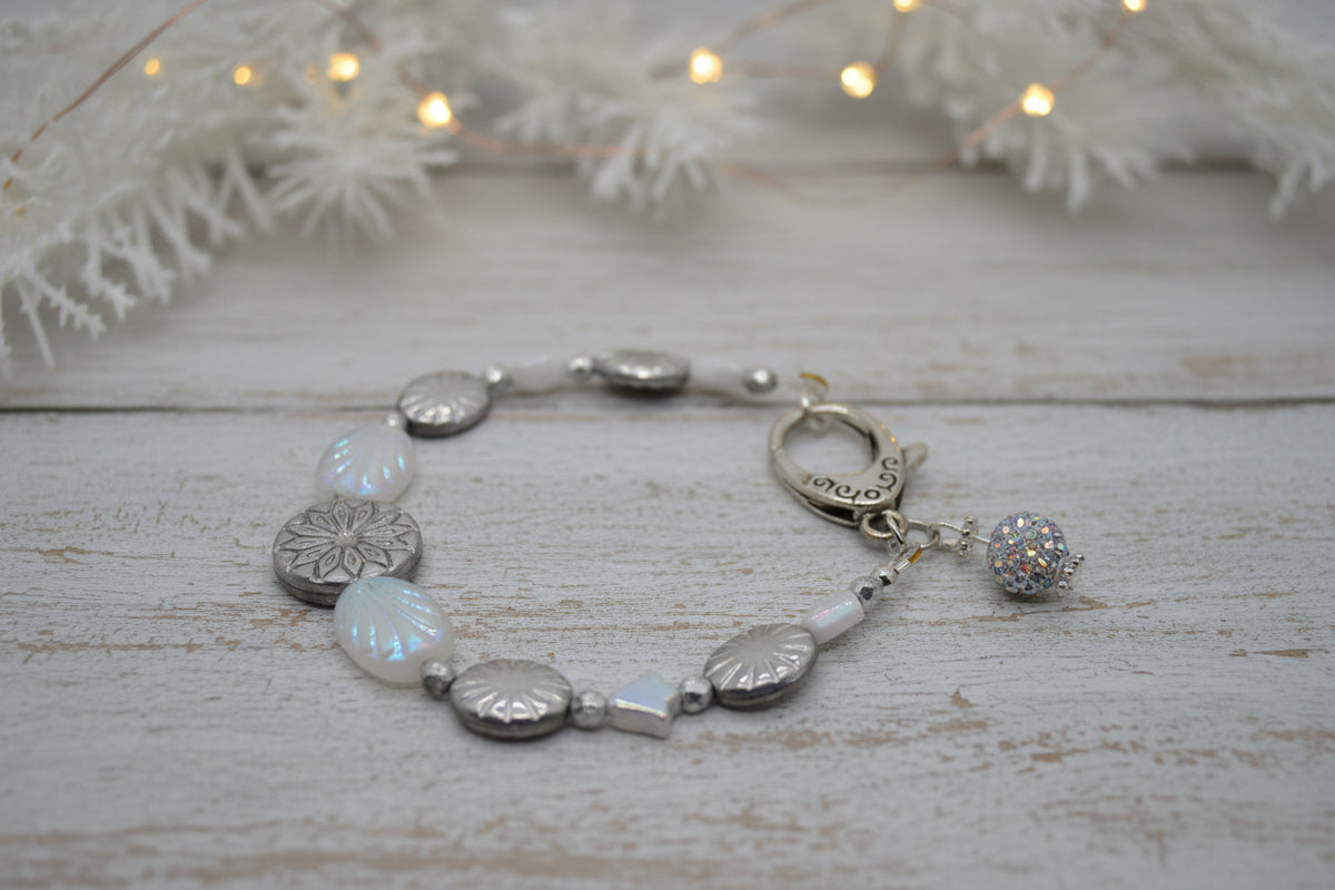 A silver-toned beaded bracelet featuring textured metallic oval beads, milky white round beads, a small iridescent star charm, and a dangling beaded ball charm, displayed on a neutral gray background.