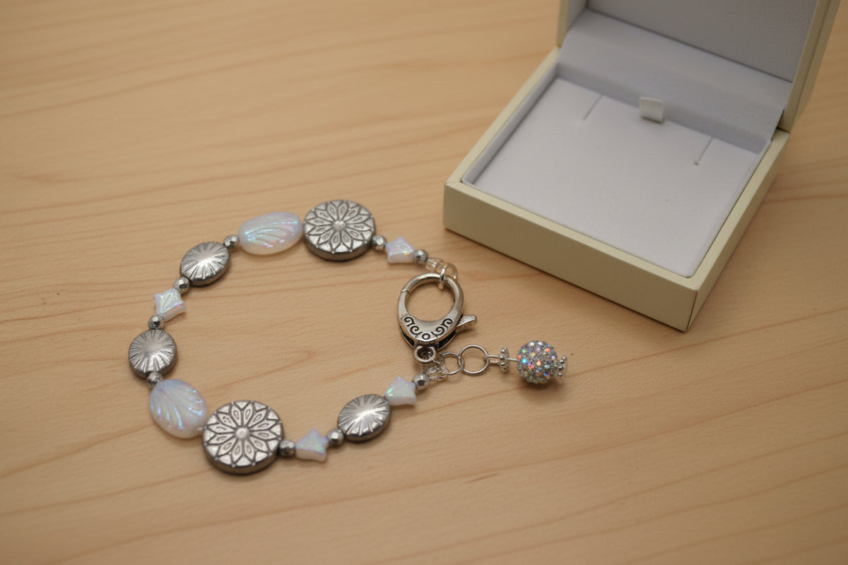 A beaded bracelet with silver-toned metal beads, iridescent white oval beads, a silver toggle clasp, and a small rhinestone ball charm, displayed on a neutral gray background.