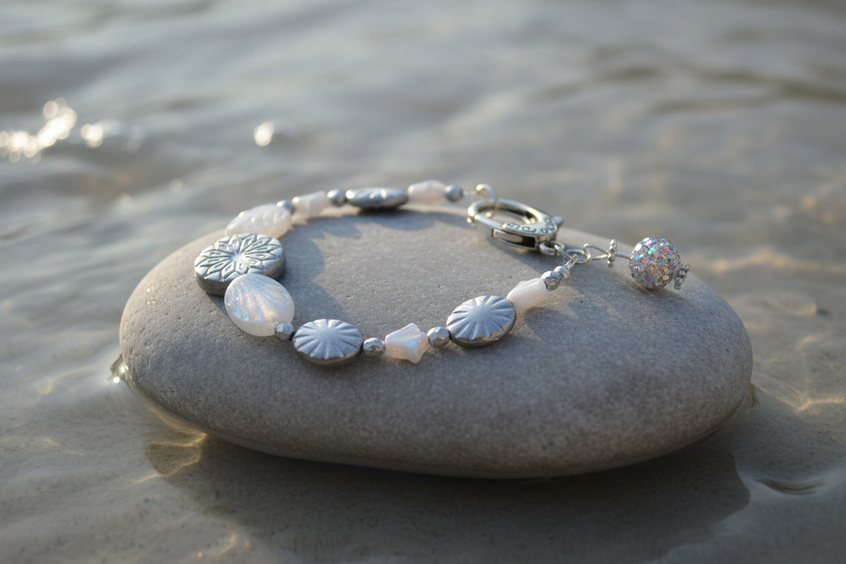 A silver-toned beaded bracelet with textured metallic oval beads, iridescent white beads, a lobster clasp, and a small colorful rhinestone ball charm, displayed on a neutral gray background.