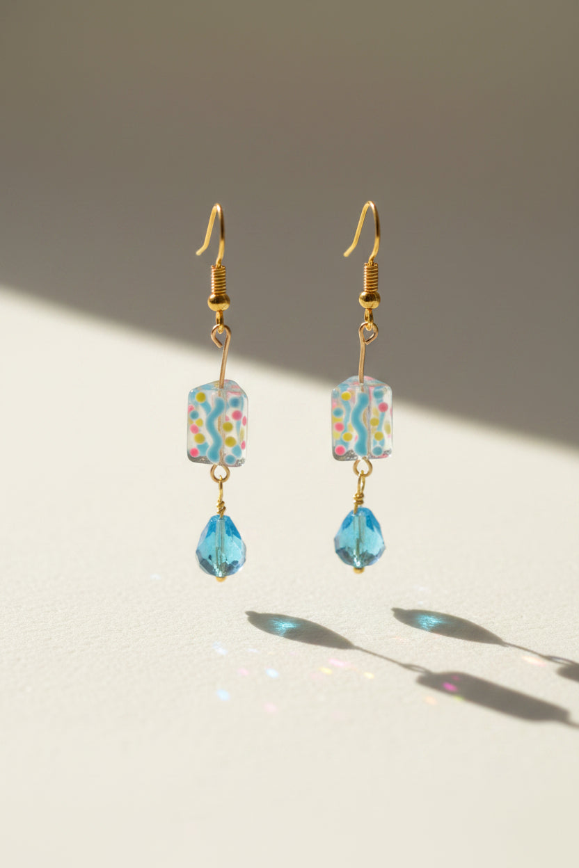 A pair of handmade dangle earrings with gold-tone hooks, a colorful patterned square bead, and a dangling light blue faceted bead, displayed on a clear acrylic stand against a light gray background.