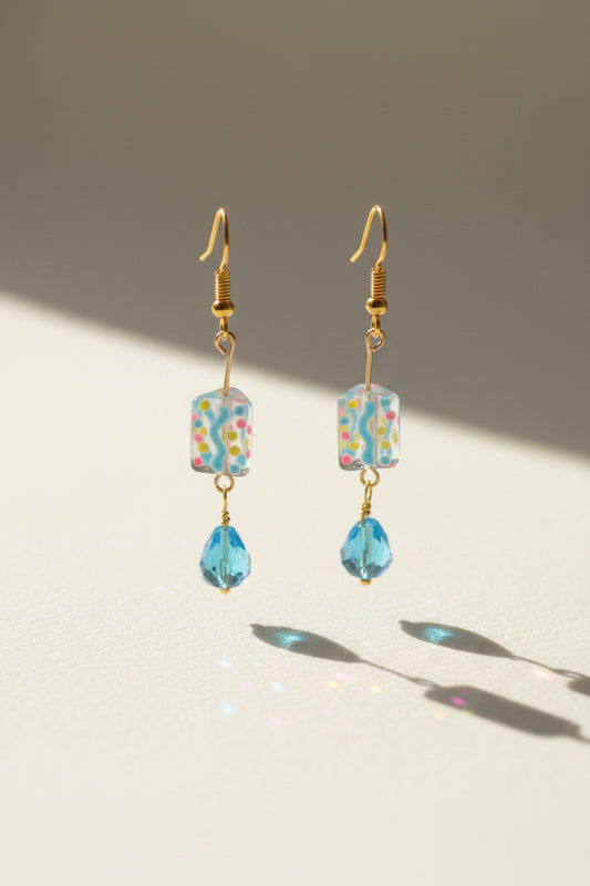 A pair of handmade dangle earrings with gold-tone hooks, a colorful patterned square bead, and a dangling light blue faceted bead, displayed on a clear acrylic stand against a light gray background.