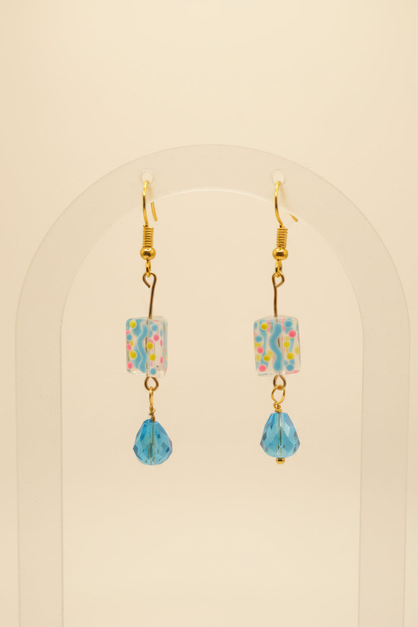 A pair of handmade dangle earrings with gold-tone hooks, square glass beads filled with colorful confetti, and light blue faceted teardrop beads hanging below, displayed on a clear acrylic stand against a neutral gray background.