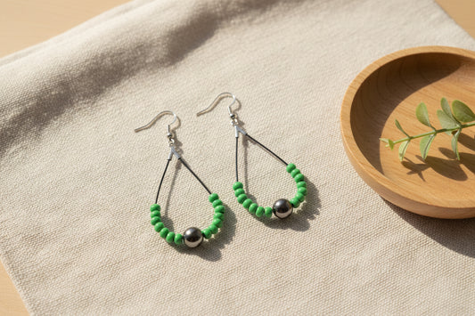 A pair of dangling earrings displayed on a clear acrylic stand against a light gray background. Each earring has a silver fishhook wire, a small silver bead, a green beaded semicircle, and a dark gray round bead at the bottom.