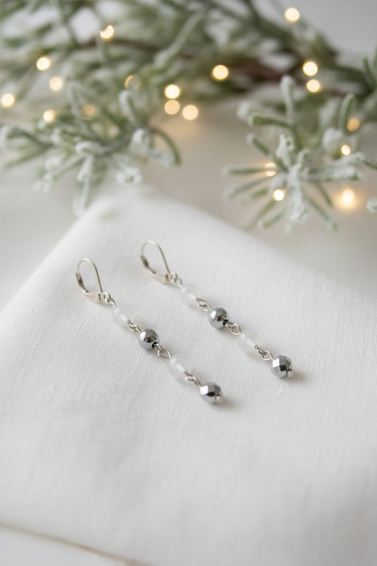 A pair of long drop earrings with small metallic beads and a dangling heart charm, displayed on a clear stand against a light gray background.