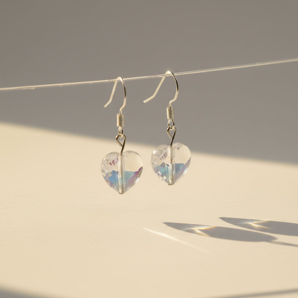 A pair of silver-toned dangle earrings with heart-shaped faceted iridescent beads, hanging from a clear acrylic display stand against a plain light gray background.