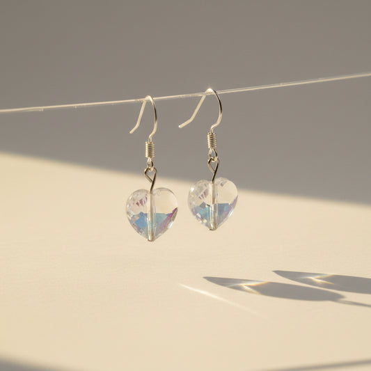 A pair of silver-toned dangle earrings with heart-shaped faceted iridescent beads, hanging from a clear acrylic display stand against a plain light gray background.