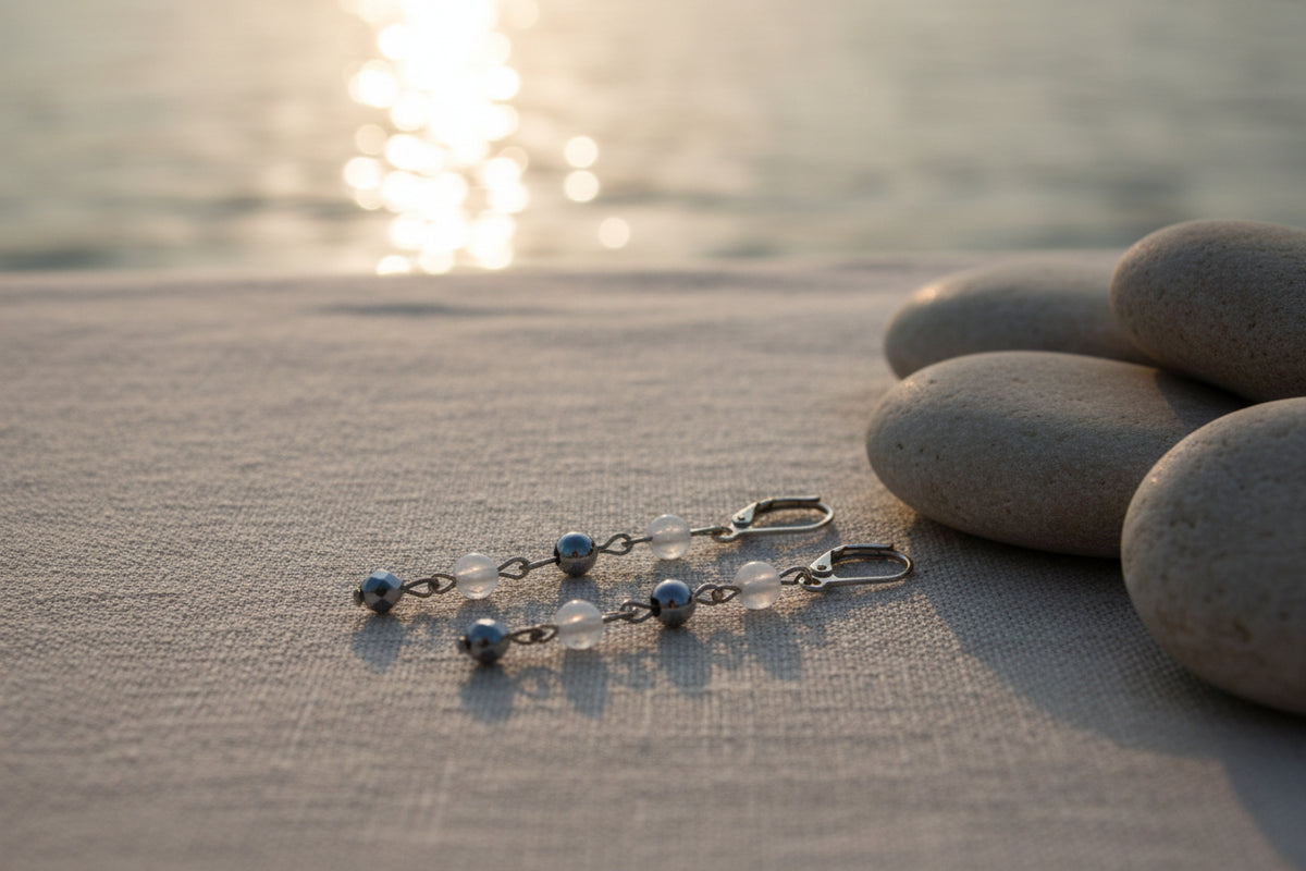 A pair of dangling earrings displayed on a clear stand against a light gray background. Each earring has a gold fishhook back, small clear beads, silver-toned chain links, a polished silver bead, and a dangling silver heart-shaped charm.