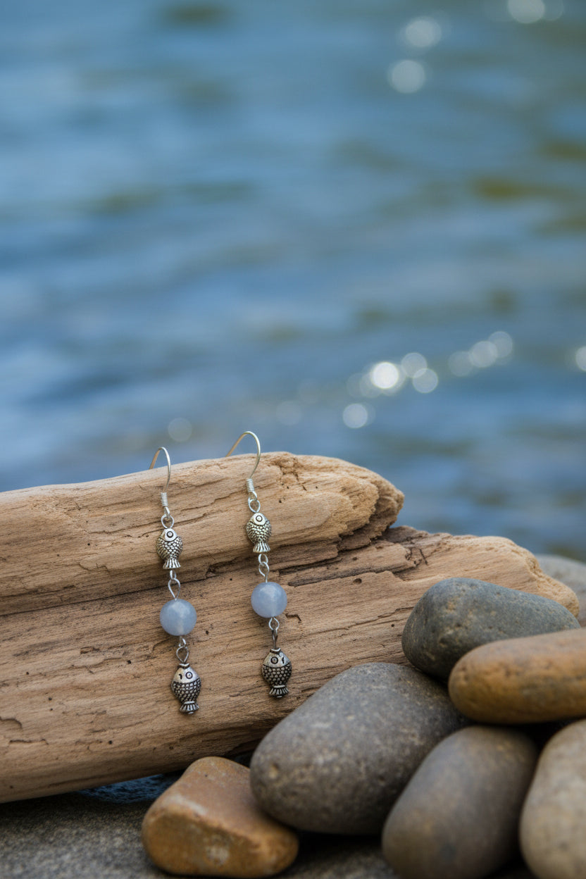 A pair of dangling earrings displayed on a clear acrylic stand against a light gray background. Each earring has a silver fishhook ear wire, a small textured metallic bead, a smooth pale blue-gray bead, and a dangling textured metallic charm.