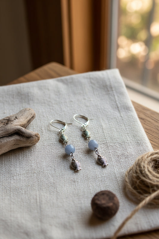 A pair of dangling earrings displayed on a clear acrylic stand against a light gray background. Each earring has a silver-toned fishhook ear wire, a small textured metallic bead, a smooth pale gray bead, and a dangling textured metallic charm.
