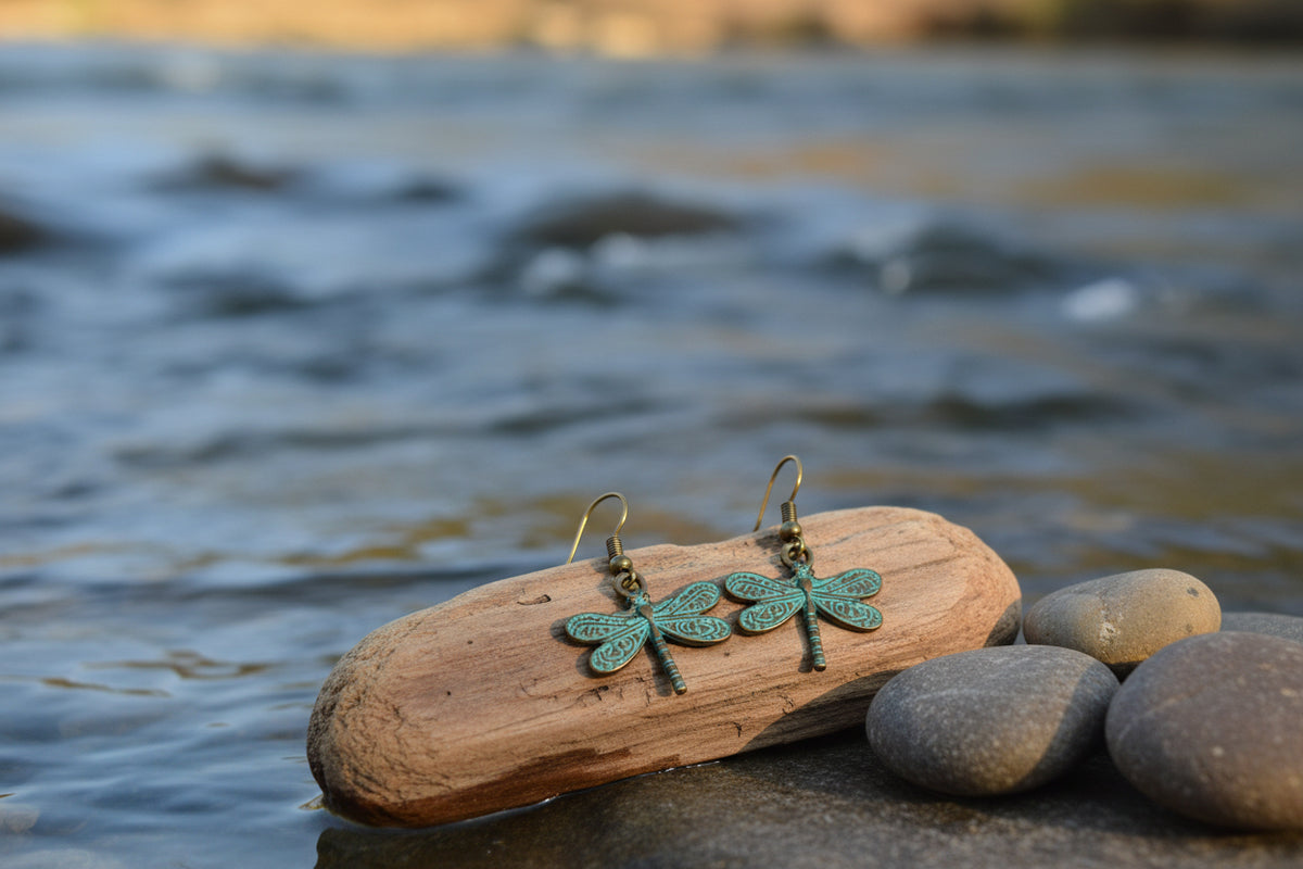 A pair of dragonfly-shaped dangle earrings with a distressed bronze finish and turquoise patina on the wings, hanging from gold-tone fishhook wires against a clear plastic display stand.