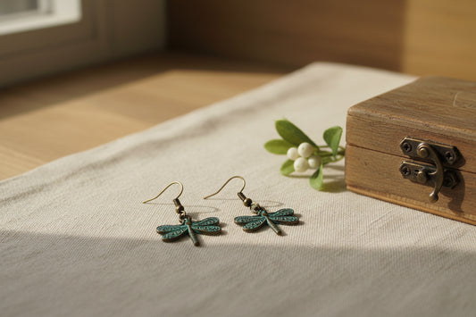 A pair of dragonfly-shaped dangling earrings with a distressed bronze finish and turquoise patina on the wings, hanging from gold-toned fishhook ear wires against a plain light gray background.