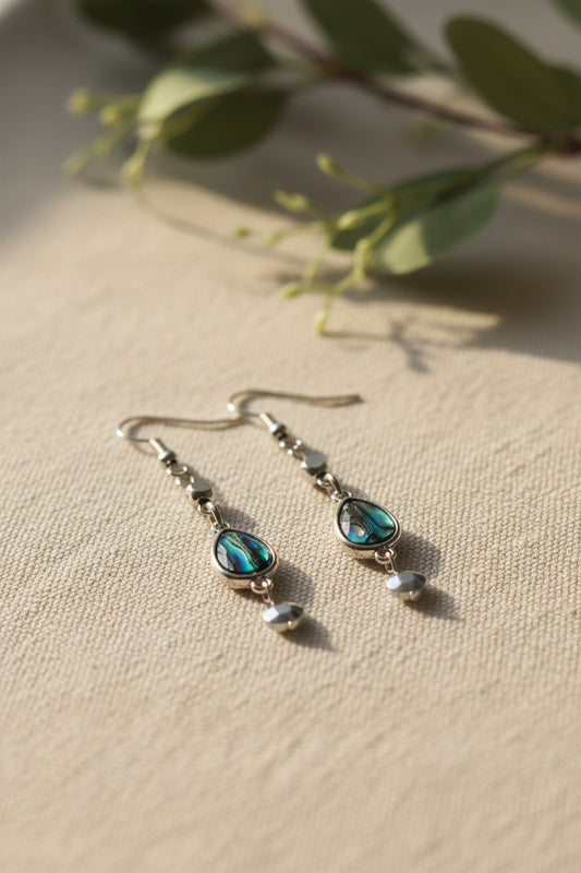 A pair of silver-toned dangle earrings with teardrop-shaped iridescent abalone shell pendants and small silver beads, displayed on a clear stand against a light gray background.