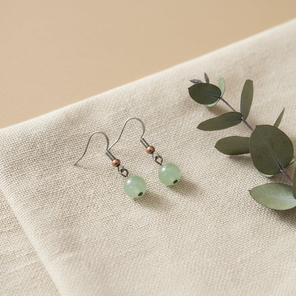 A pair of handmade dangle earrings with light green round stone beads, copper-colored spacer beads, and silver-toned hooks, displayed on a clear stand against a light gray background.