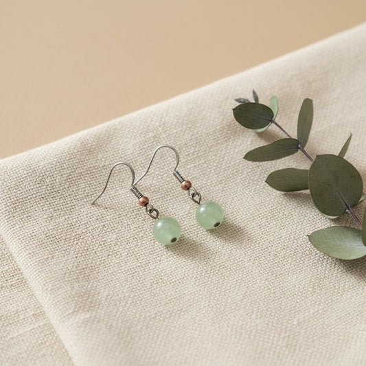 A pair of handmade dangle earrings with light green round stone beads, copper-colored spacer beads, and silver-toned hooks, displayed on a clear stand against a light gray background.