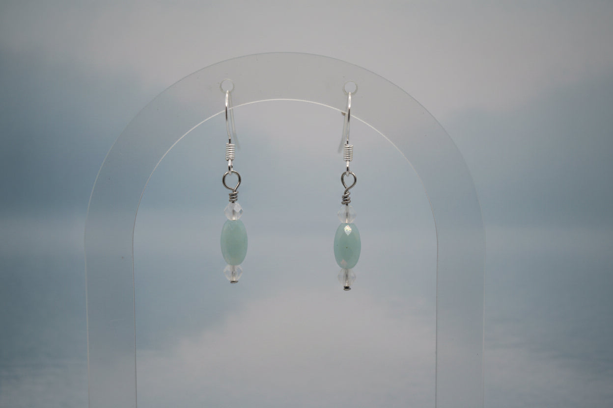 A pair of dangling earrings with oval light blue beads and silver hooks, displayed on a clear acrylic stand against a soft gray background.