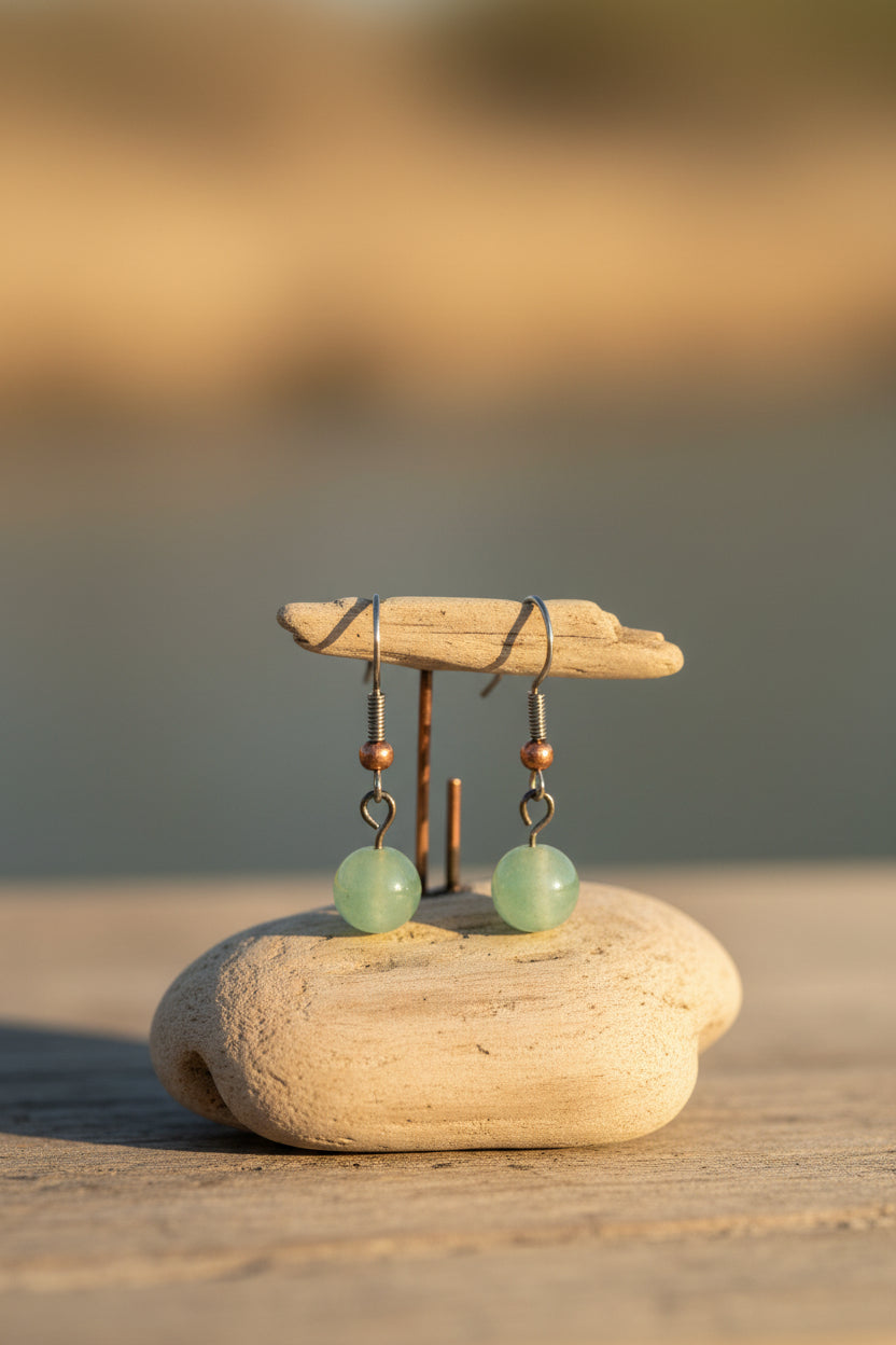 A pair of handmade dangle earrings featuring light green jadeite round beads, copper-colored spacer beads, and silver-toned metal hooks, displayed on a clear plastic stand against a neutral gray background.