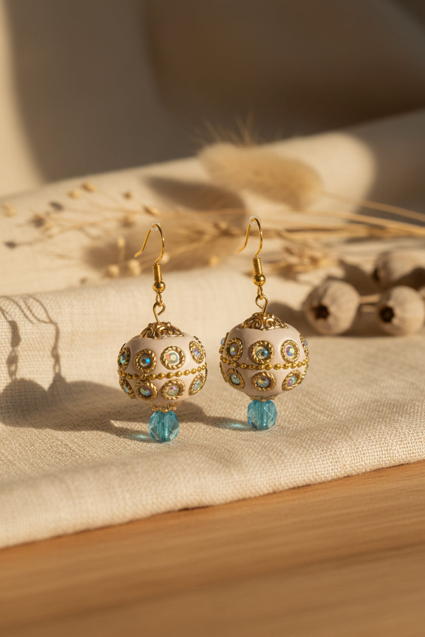 A pair of ornate gold-tone dangle earrings with spherical cream-colored beads decorated with small blue and green stones, hanging from gold fishhook wires. The earrings are displayed against a neutral gray background with a subtle arch.