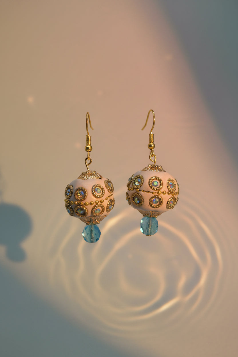 A pair of ornate gold-tone dangle earrings with cream-colored spherical beads, decorated with small blue and green rhinestones, and finished with dangling light blue beads at the bottom, displayed against a neutral gray background.