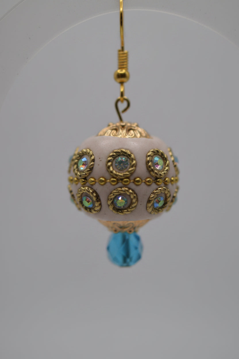A pair of handmade dangle earrings with cream-colored glass beads adorned with gold filigree and iridescent rhinestones, hanging from gold hooks with light blue bead drops.