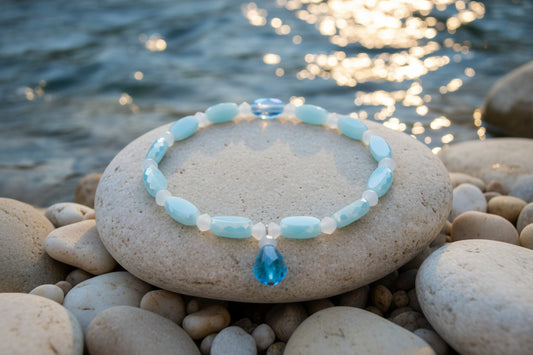 A beaded stretch bracelet featuring light blue and white beads, with one faceted light blue crystal charm hanging from the front, displayed on a neutral light gray background.