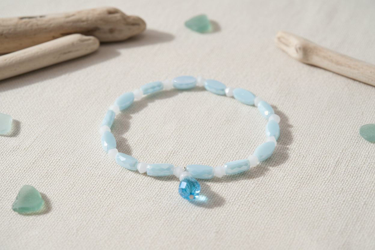A beaded bracelet featuring light blue and white beads, including iridescent oval beads and a faceted blue bead charm, displayed on a plain light gray background.