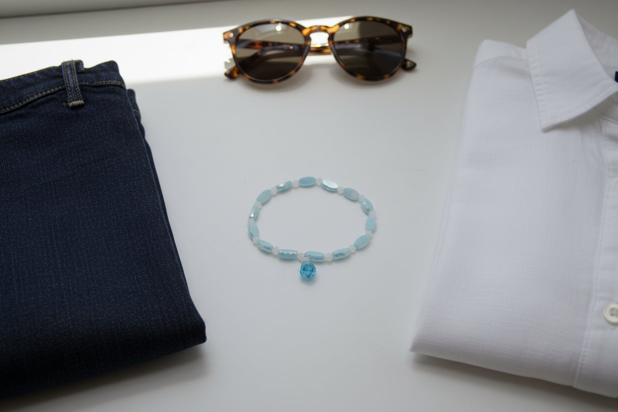 A beaded stretch bracelet featuring light blue oval beads, translucent white round beads, and a small dangling light blue bead charm, displayed on a plain light gray background.