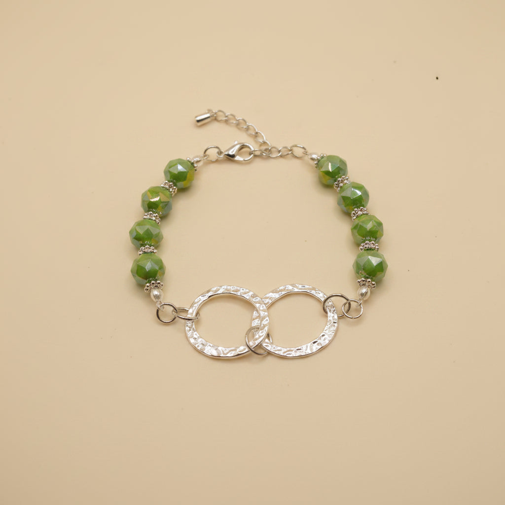 A beaded bracelet featuring green marbled faceted beads and a central silver-tone interlocking double ring design with clear rhinestone-like accents, shown on a plain light gray background.