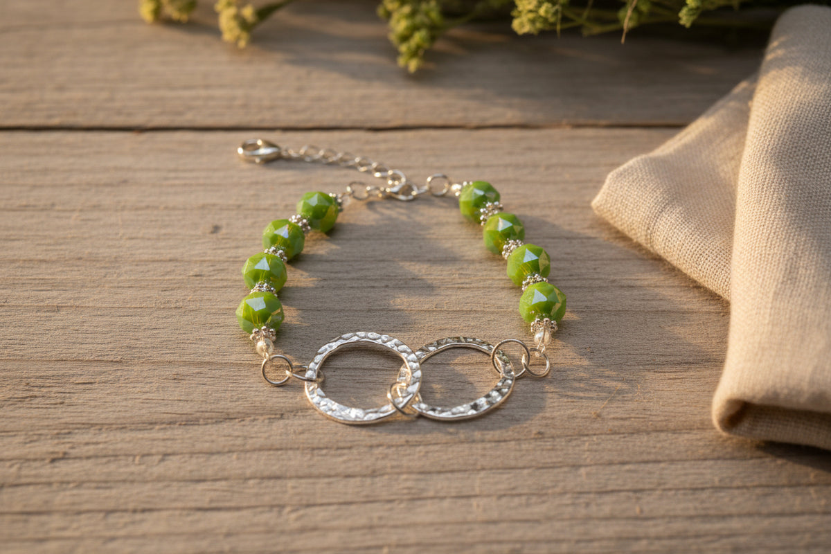 A beaded bracelet featuring green faceted beads and a silver-tone double interlocking ring charm, displayed on a plain light gray surface.