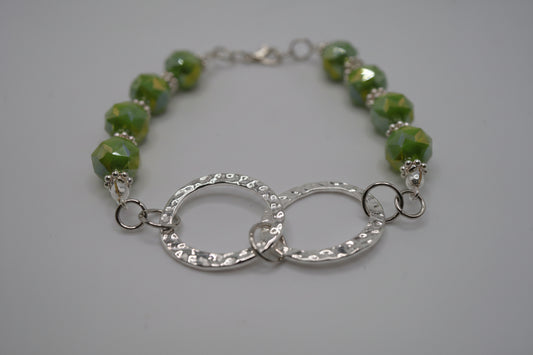 A beaded bracelet featuring green marbled beads and a textured silver double-ring centerpiece, displayed on a neutral gray background.
