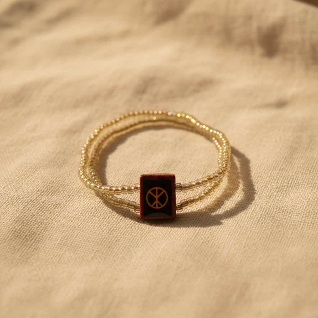 A beaded wrap bracelet with a central square charm featuring a black background, a gold peace sign, and a red border, displayed on a plain light gray surface.