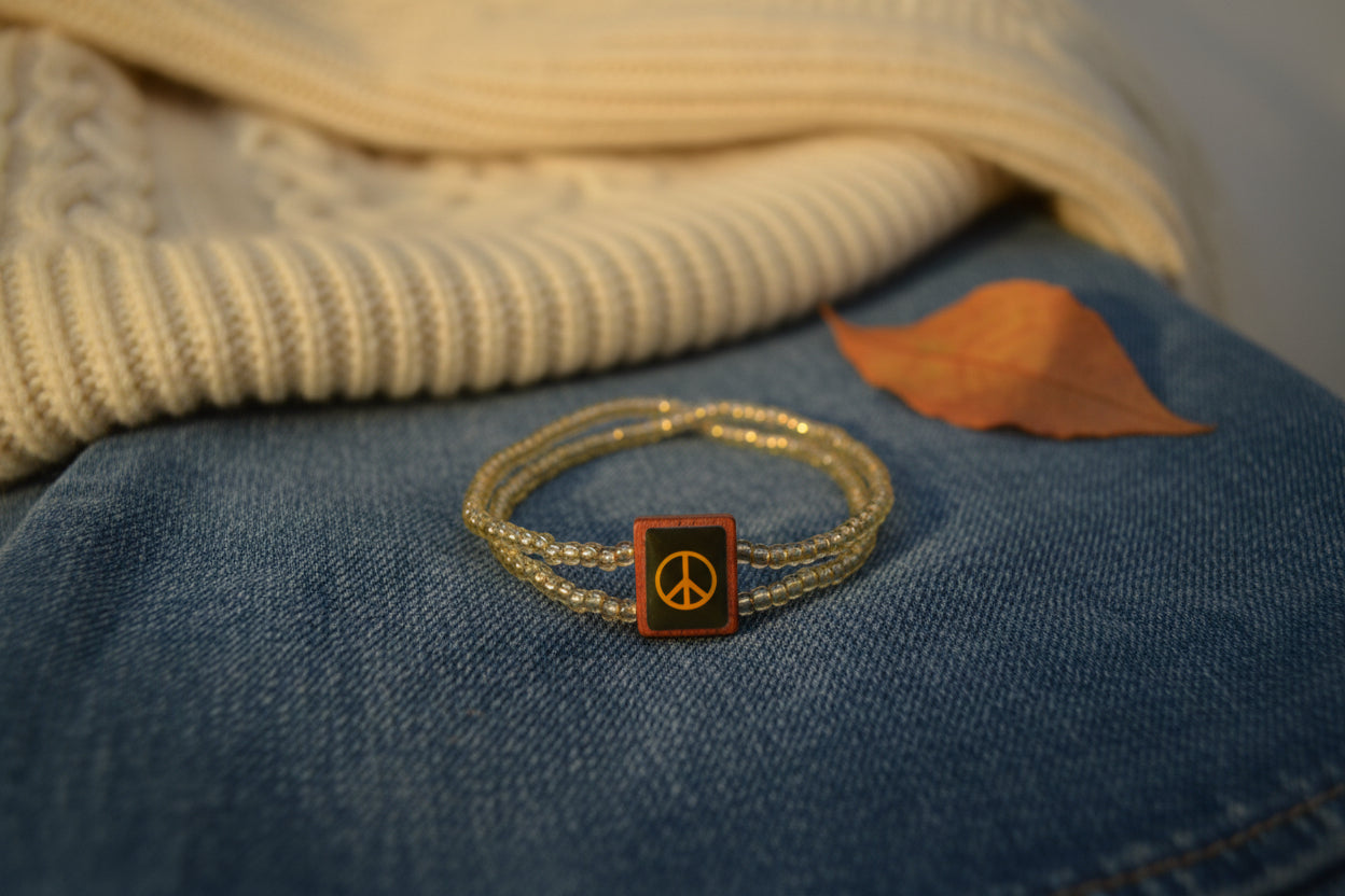 A beaded wrap bracelet with a central square charm featuring a black background, a yellow stylized symbol, and a small red accent, displayed on a plain white surface.