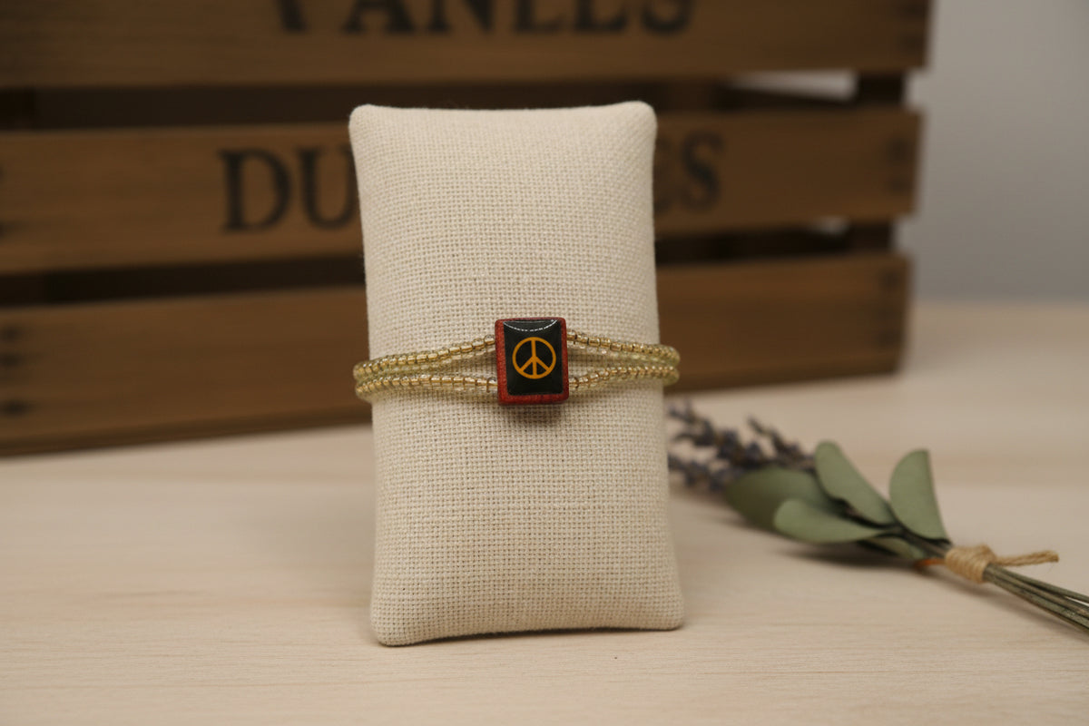 A beaded wrap bracelet with a central square charm featuring a yellow symbol on a black background, set against a plain light gray backdrop.