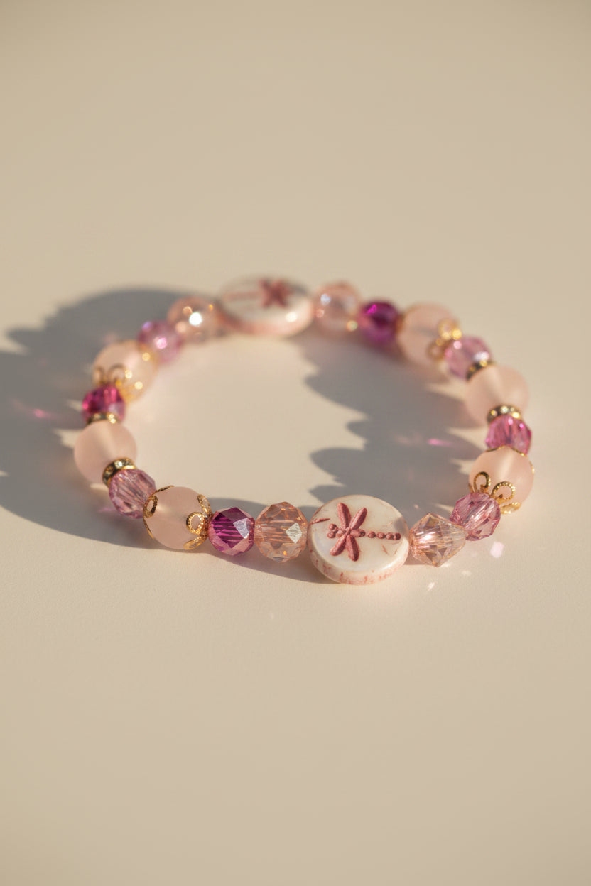 A beaded stretch bracelet featuring pink, peach, and white beads with metallic gold-toned details, including two round white beads decorated with pink starburst designs, displayed on a neutral gray background.