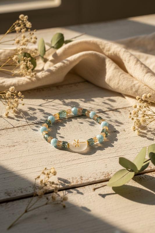 A beaded stretch bracelet featuring light blue round beads, faceted dark green and gold beads, and a central round white bead with a gold symbol, displayed on a neutral gray background.
