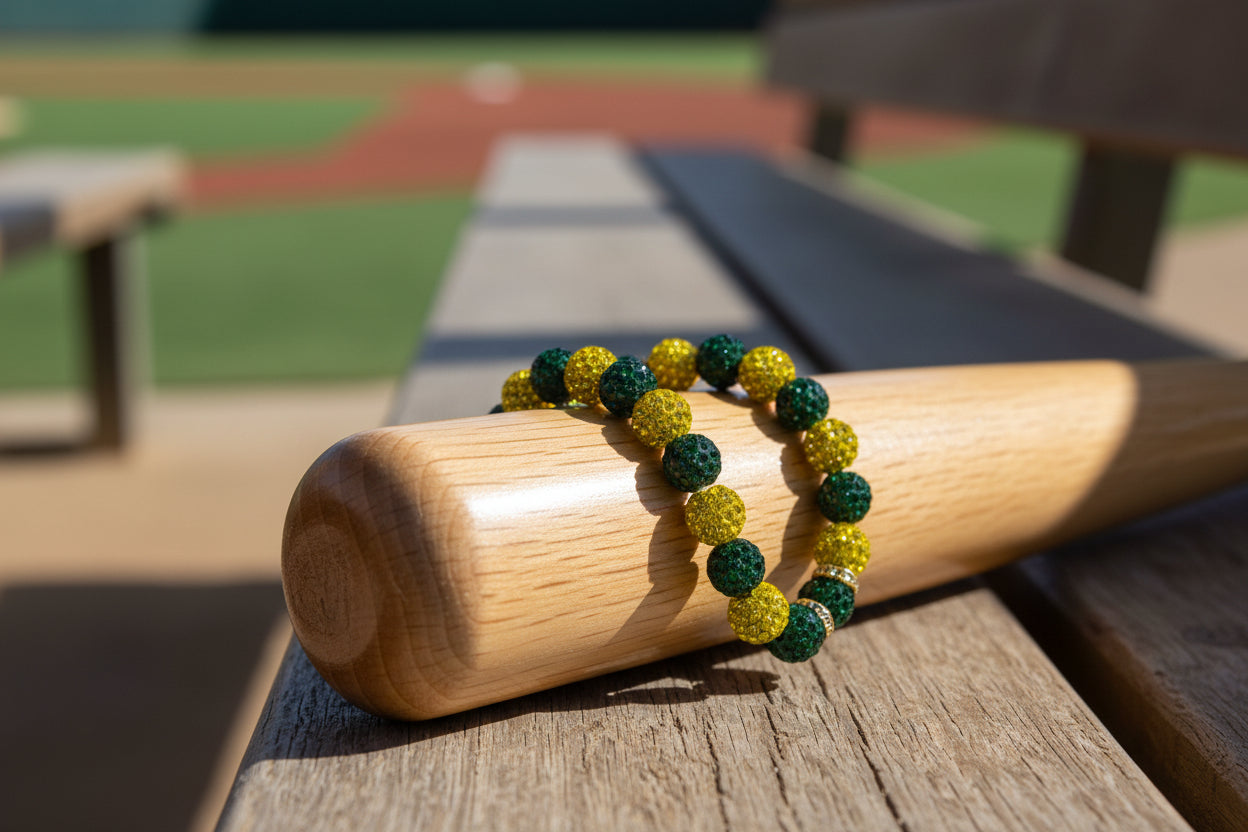 A beaded stretch bracelet with green and yellow textured beads rests on a wooden baseball bat, set on a wooden bench with a blurred baseball field in the background.