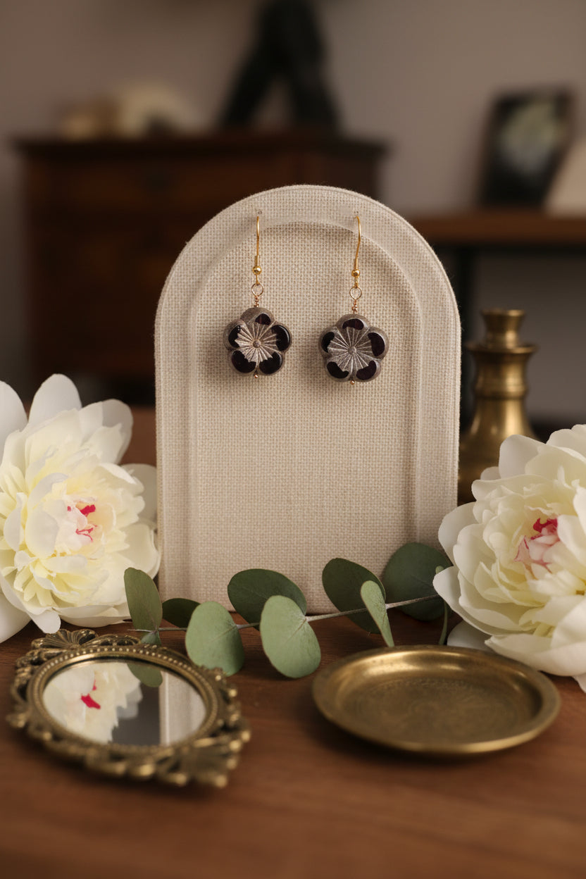 A pair of flower-shaped drop earrings with dark purple petals, textured gray centers, and gold fishhook wires, displayed against a light gray background.