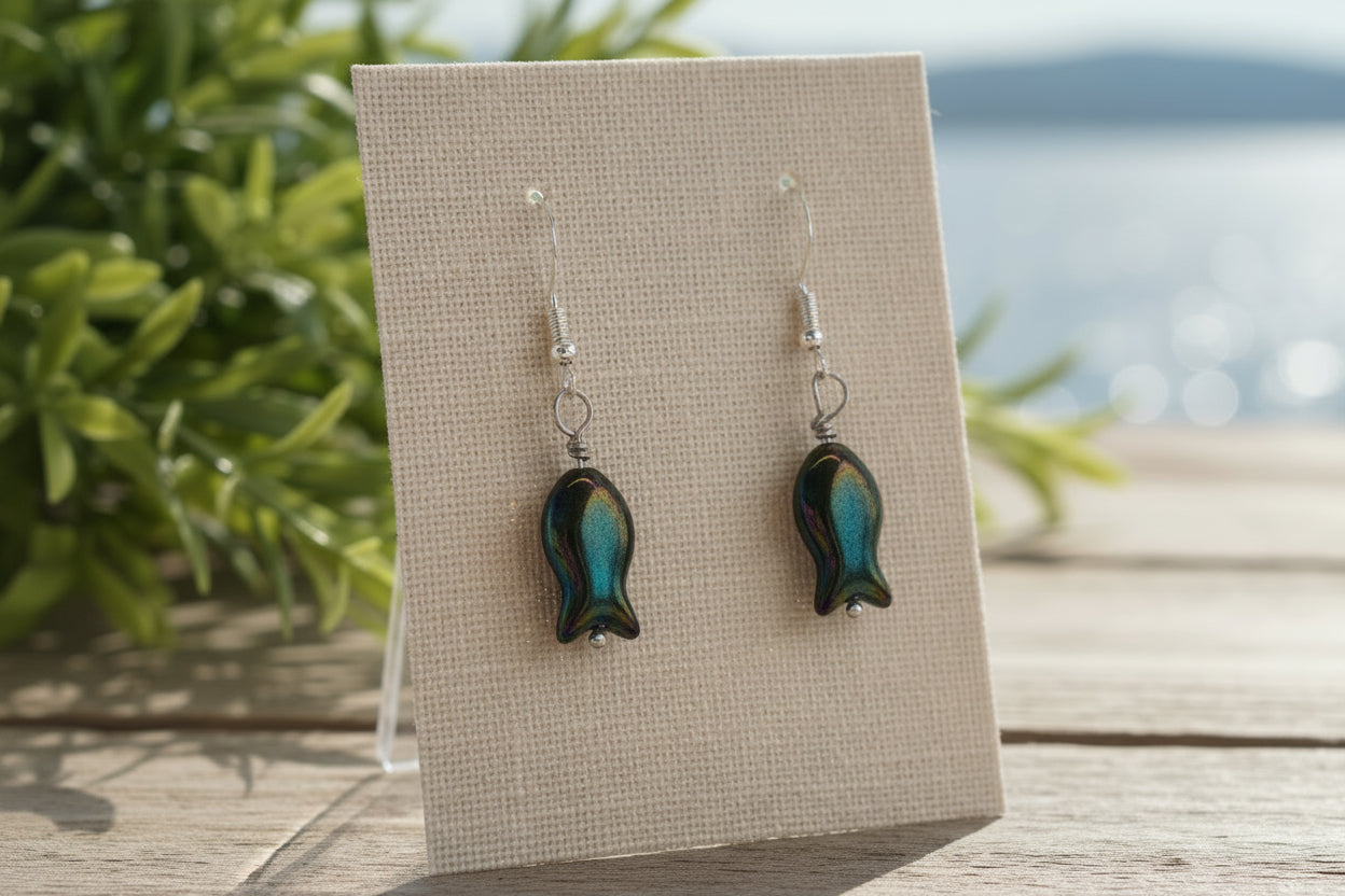 A pair of fish-shaped drop earrings with iridescent dark green and blue glass bodies and silver fishhook wires, displayed on a clear plastic stand against a neutral gray background.